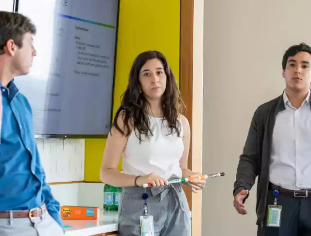 Three young professionals stand in a modern office, one woman holding a marker and presenting near a large screen displaying a presentation. The other two men listen attentively. Office supplies and water bottles are visible on the counter.
